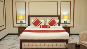 Una habitación de hotel con una cama grande, dos mesitas de noche con lámparas, un cabecero con almohadones decorativos y un suelo de baldosas de mármol.