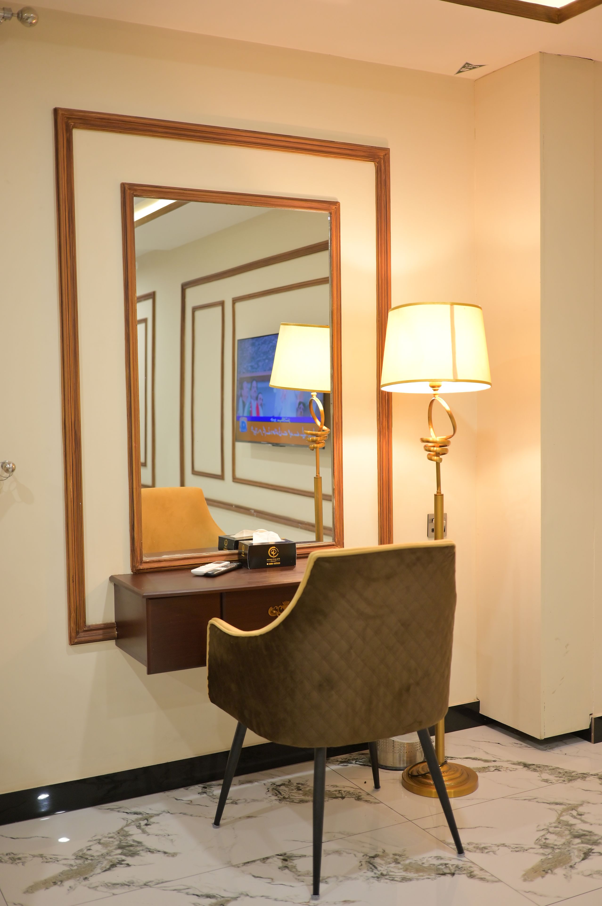 A hotel room with a large mirror, a desk with a lamp, a chair, and a television.