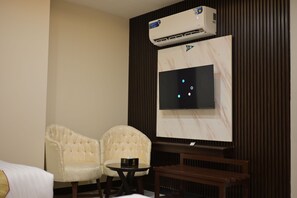 A hotel room with a bed, two armchairs, a small table, a TV mounted on the wall, and an air conditioning unit.