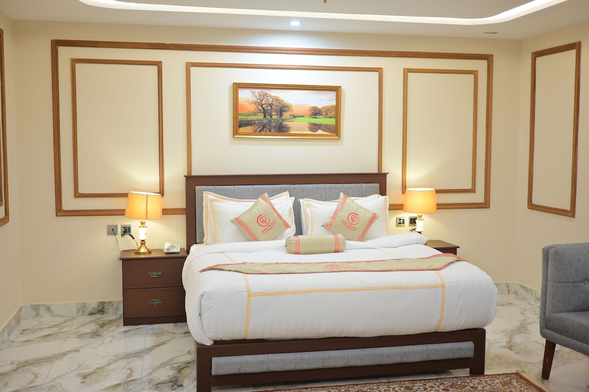 Habitación de hotel con cama, dos mesitas de noche, una lámpara, un cuadro y una silla.