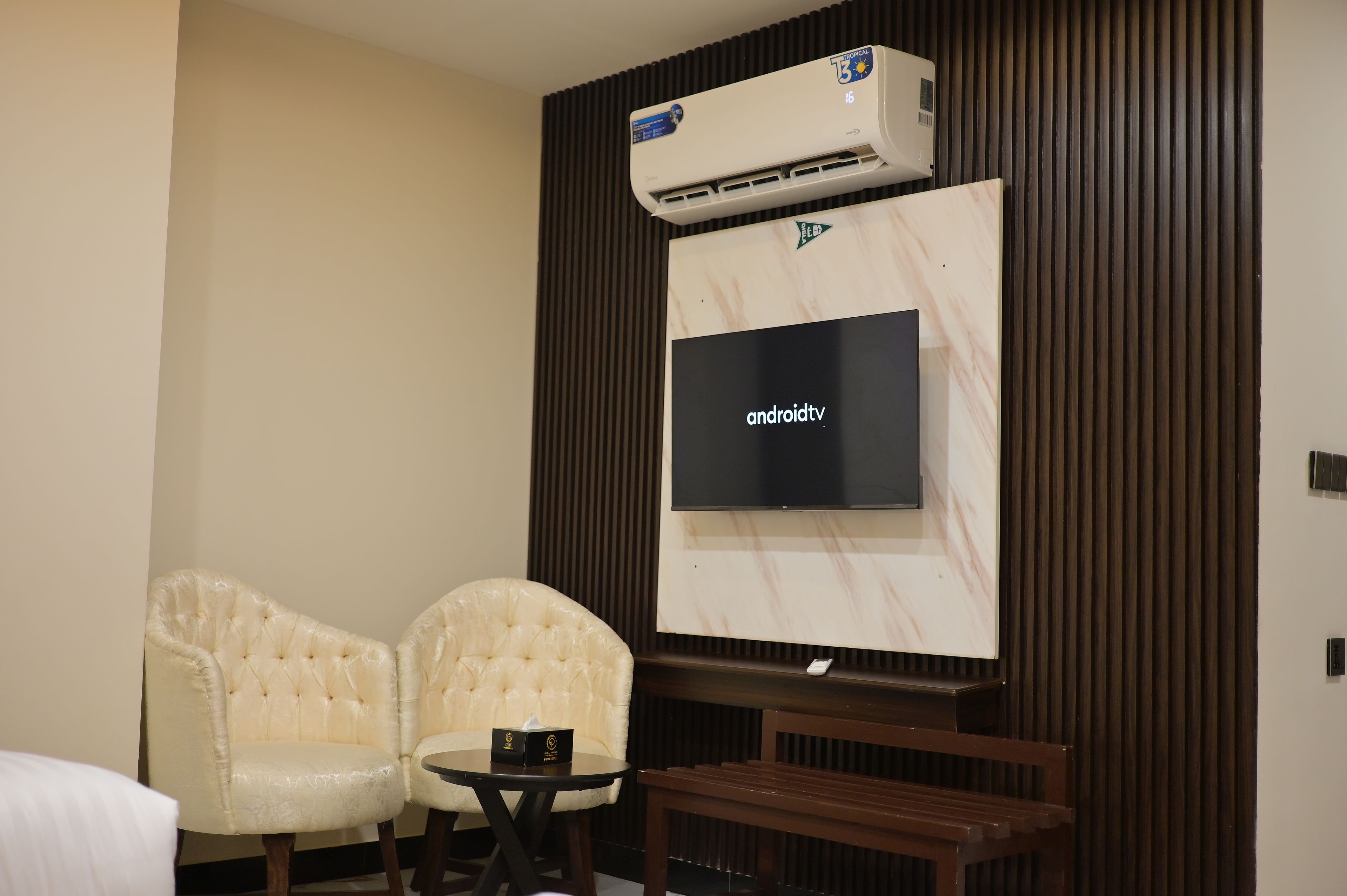 A room with a flat-screen TV mounted on a wall, two upholstered chairs, a small round table with a remote control, and an air conditioning unit above the TV.