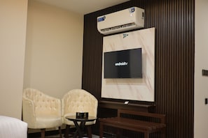 Una habitación con televisión de pantalla plana montada en la pared, dos sillones tapizados, una mesita redonda con un control remoto y un equipo de aire acondicionado sobre la televisión.