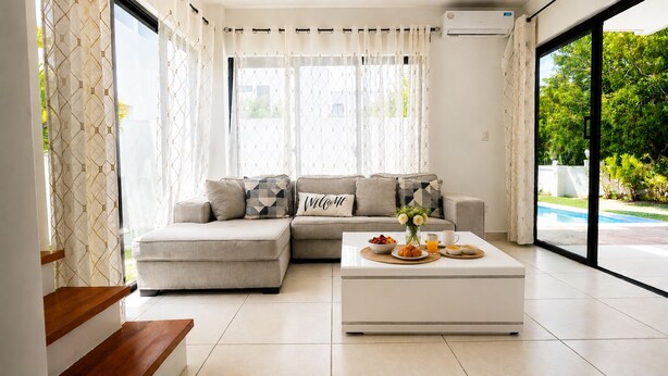 Een moderne woonkamer met een hoekbank, een bijzettafeltje met ontbijtartikelen en grote ramen met gordijnen.