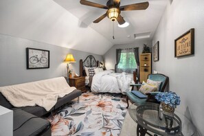 A bedroom with a bed, a sofa, a chair, a glass table, and a ceiling fan.