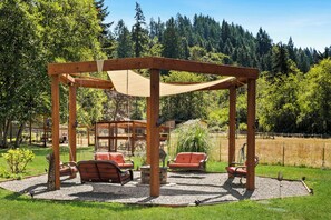 A wooden gazebo with a swing set and seating area, surrounded by a grassy field and trees.