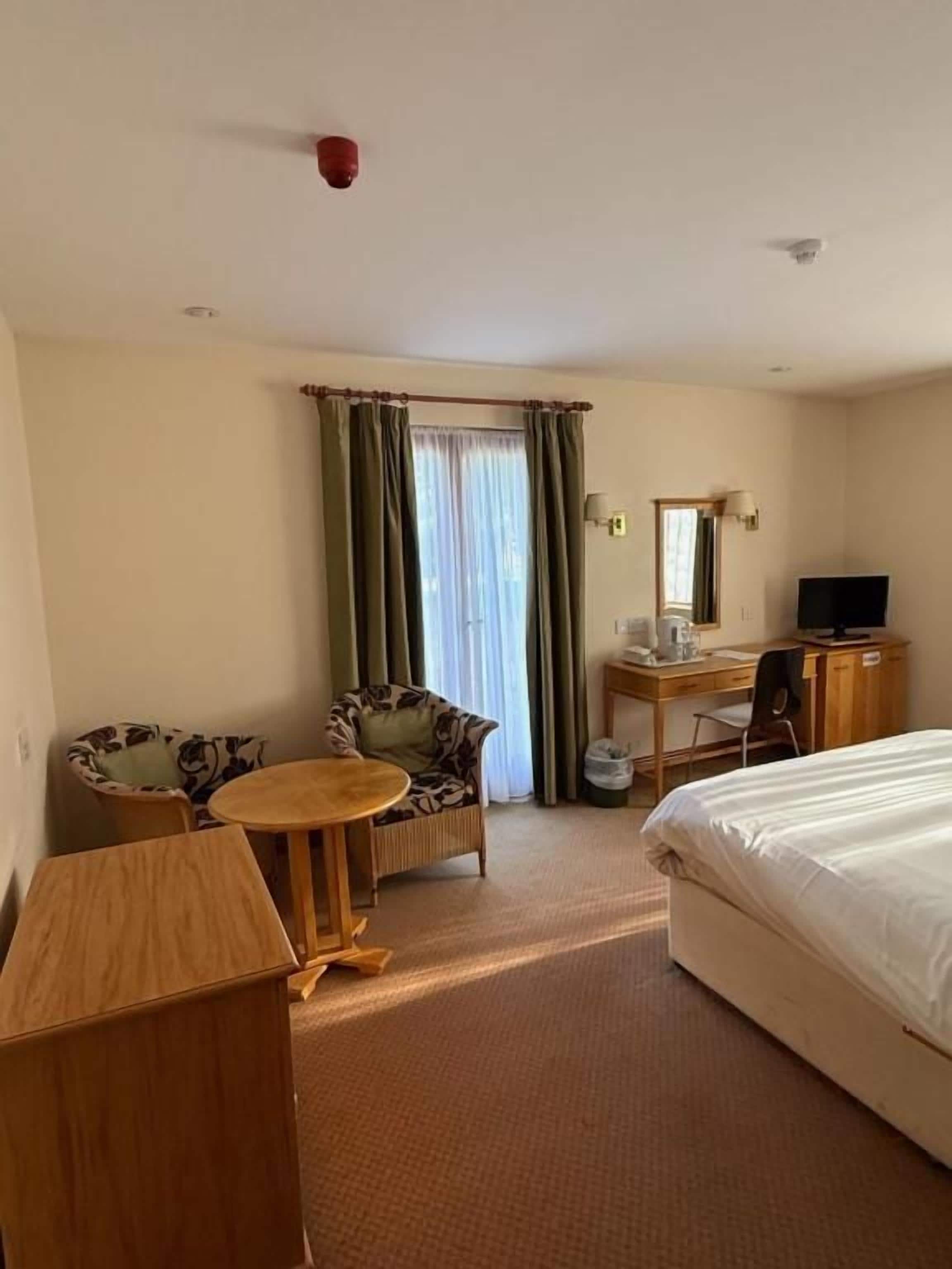A hotel room with a bed, a desk, a chair, a small table, and a window with curtains.