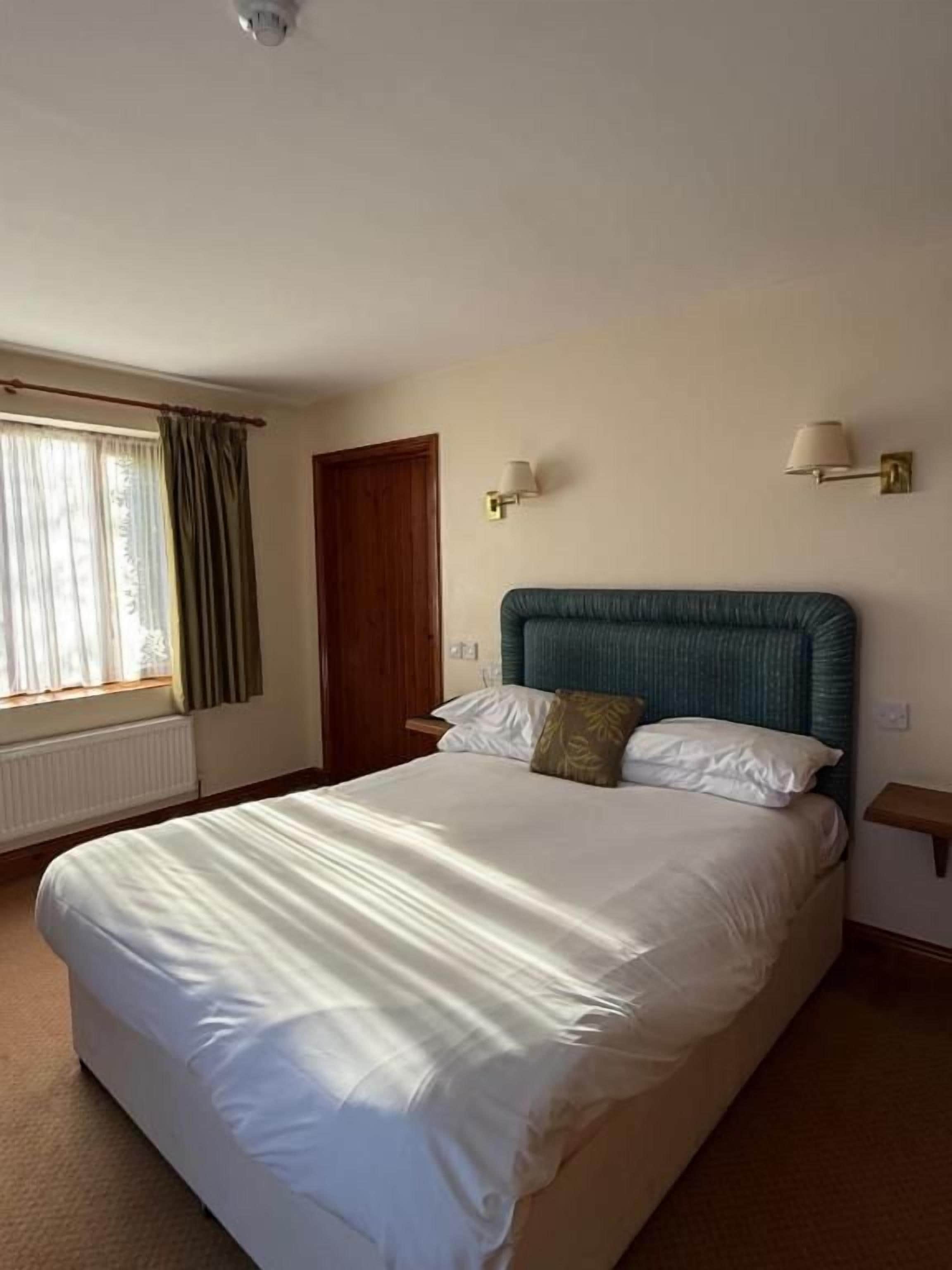 A hotel room with a large bed, a window with curtains, a door, and two wall-mounted lamps.
