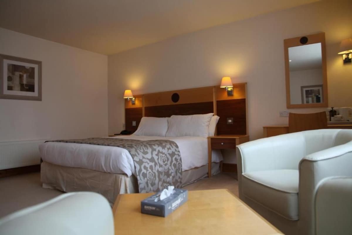Una habitación de hotel con una cama, mesitas de noche, una silla, una pequeña mesa con una caja de pañuelos y un espejo en la pared.