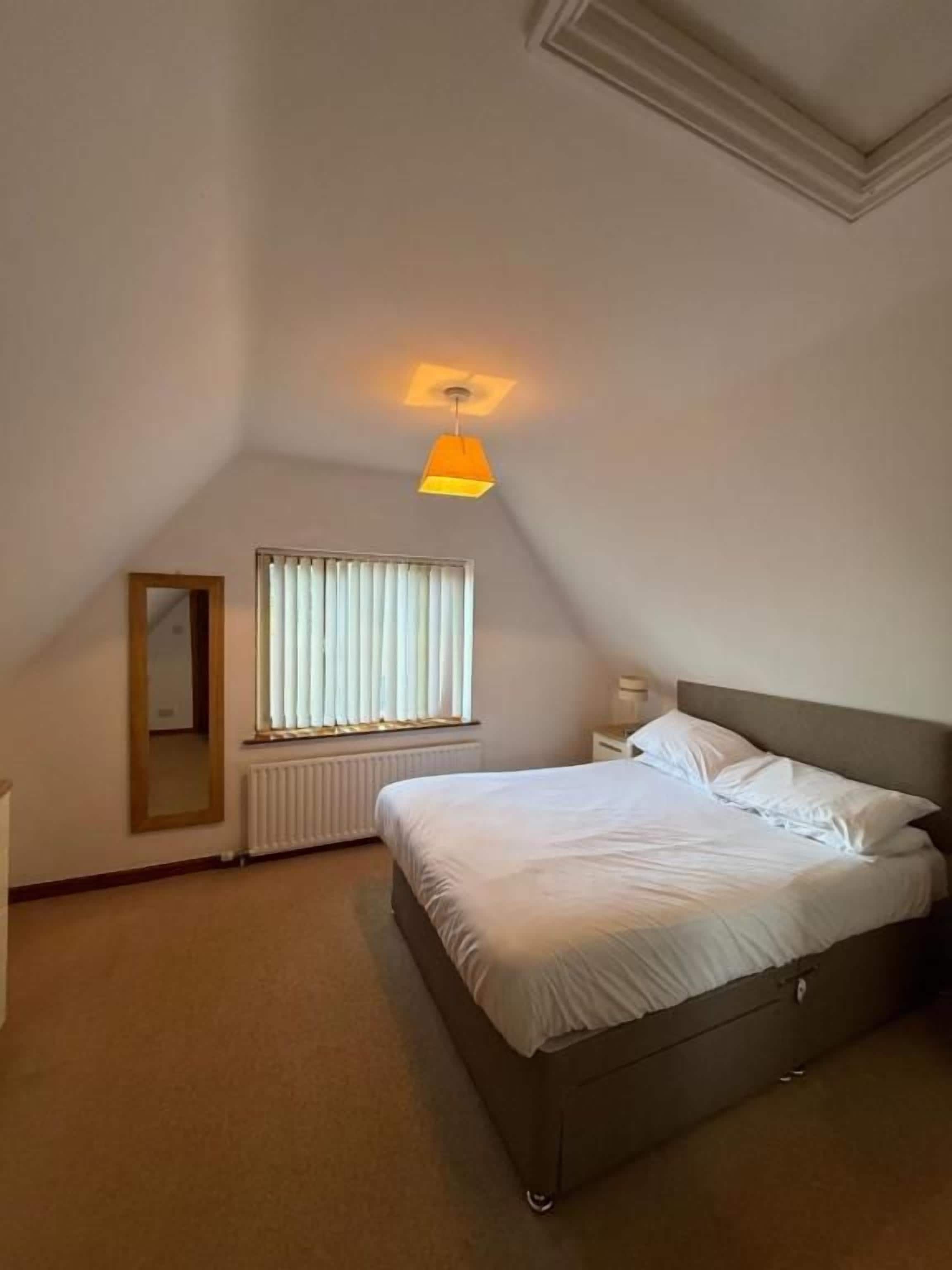 A bedroom with a bed, a bedside table, a window with blinds, a radiator, and a hanging light fixture.