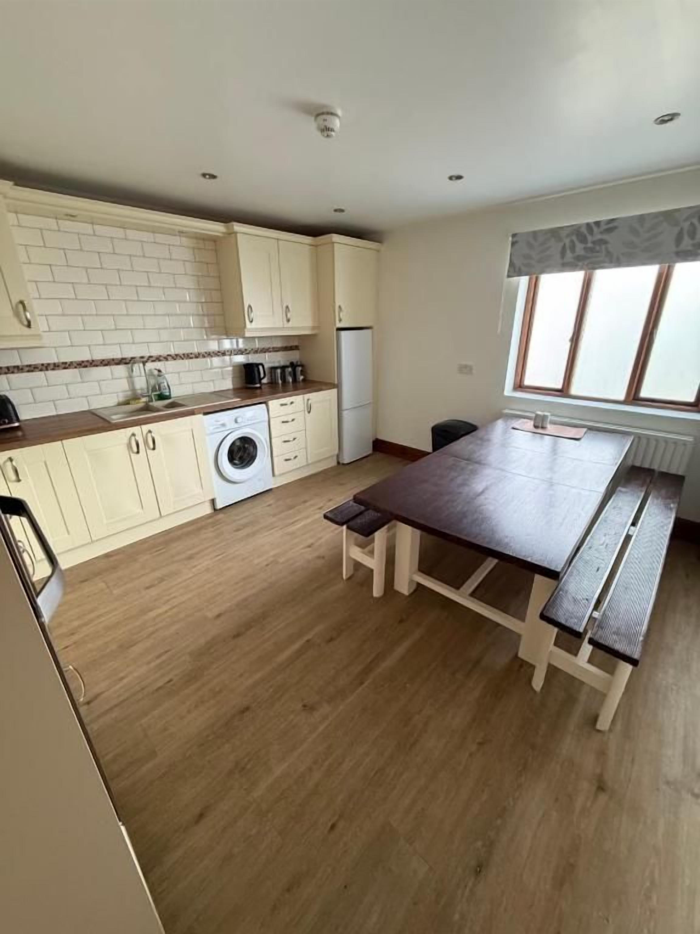 Una cocina con suelo de madera, una mesa de comedor con bancos y una ventana con persianas.