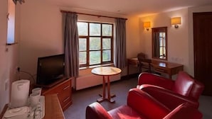 Una habitación de hotel con un sofá rojo, una mesita redonda, un televisor y una ventana con cortinas.