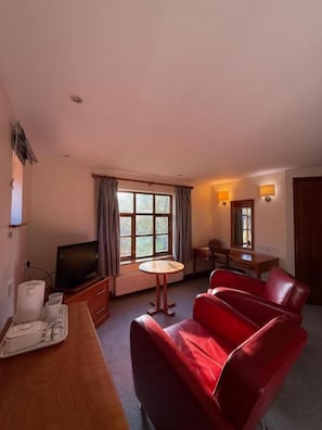Una habitación de hotel con un sofá rojo, una mesita redonda, un televisor y una ventana con cortinas.