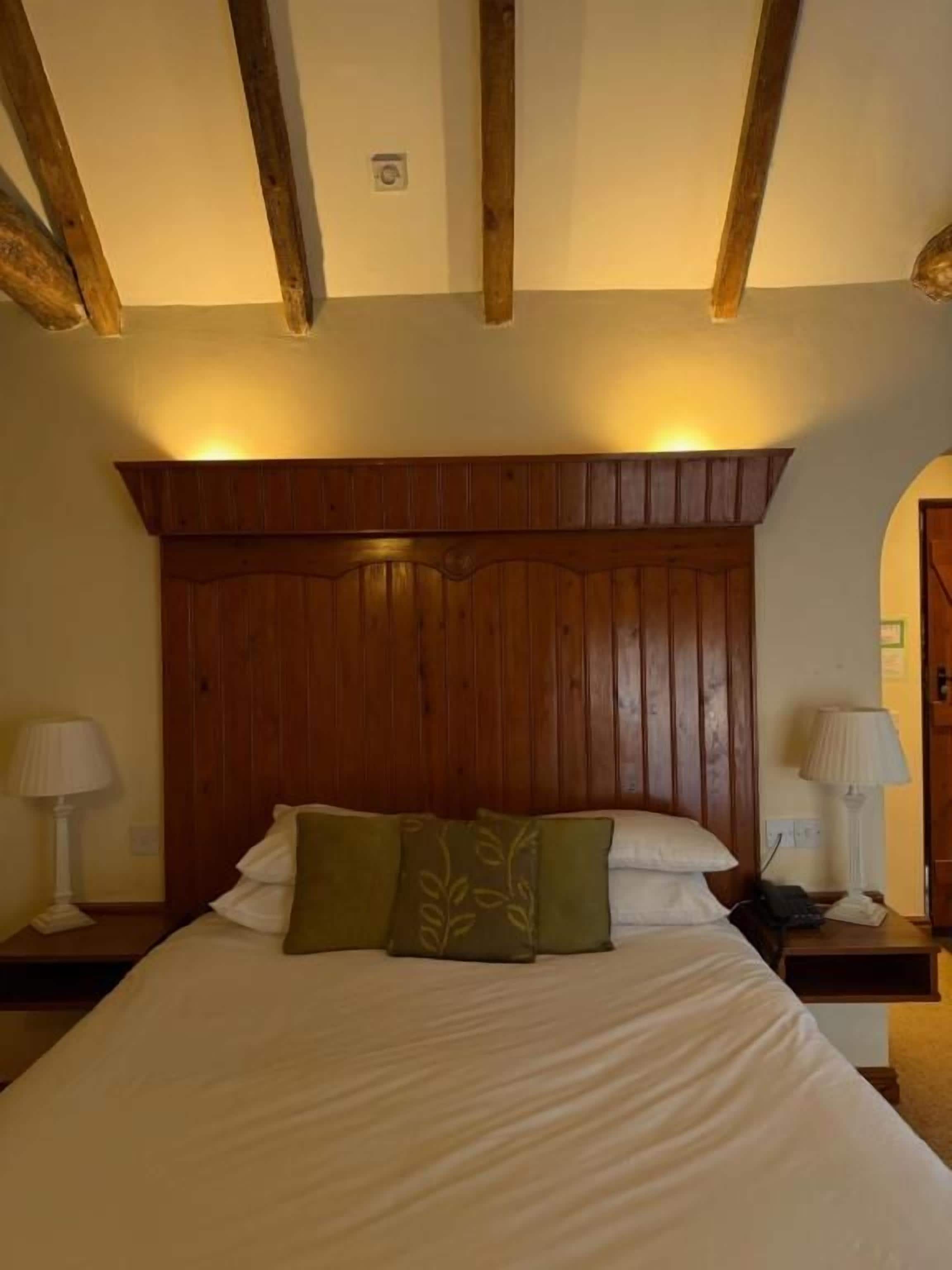 Un dormitorio con cabecera de madera, cama con ropa de cama blanca, dos mesitas de noche con lámparas y estructura de techo de madera.