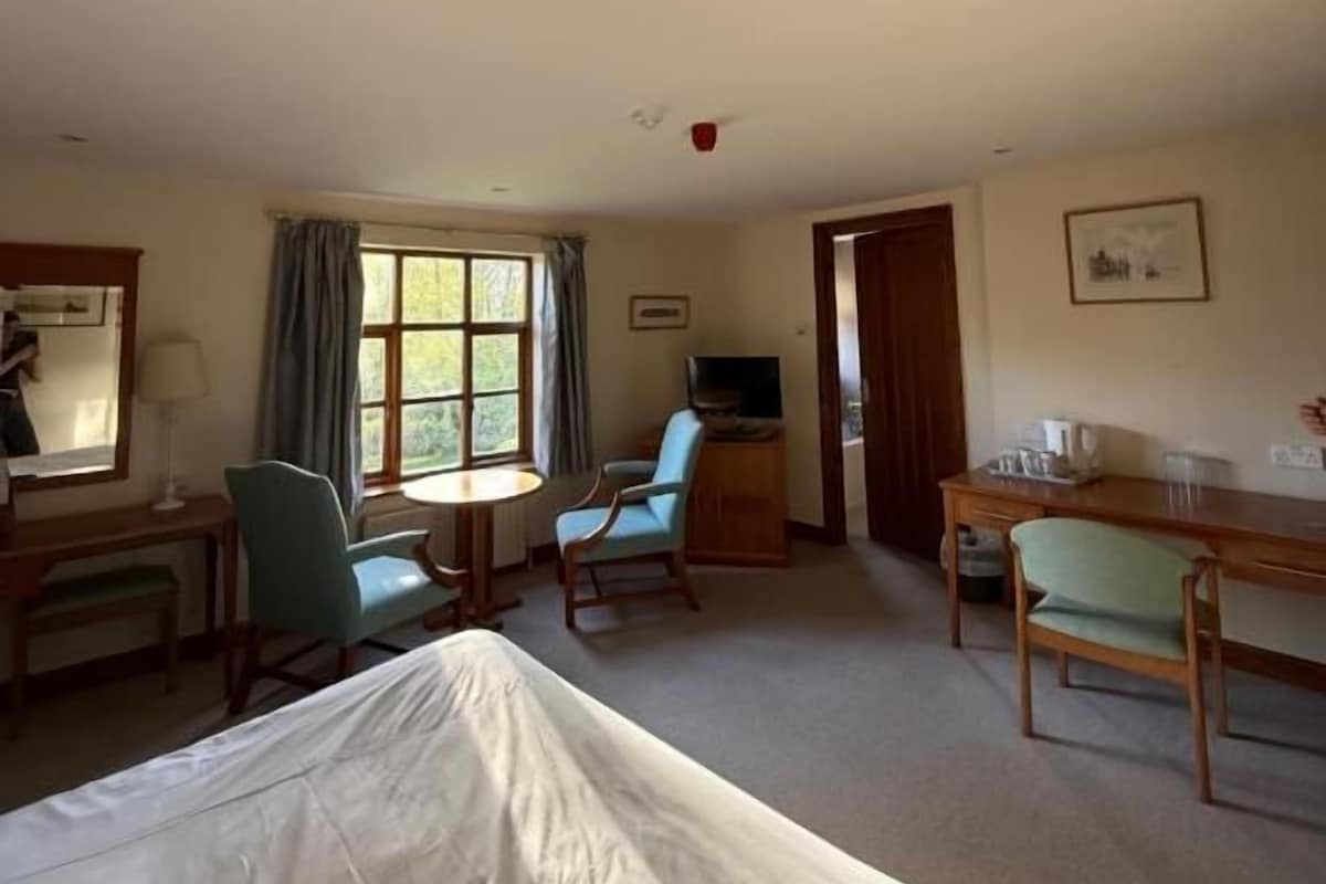 Habitación de hotel con cama, escritorio, silla, mesita, televisión y ventana con cortinas.