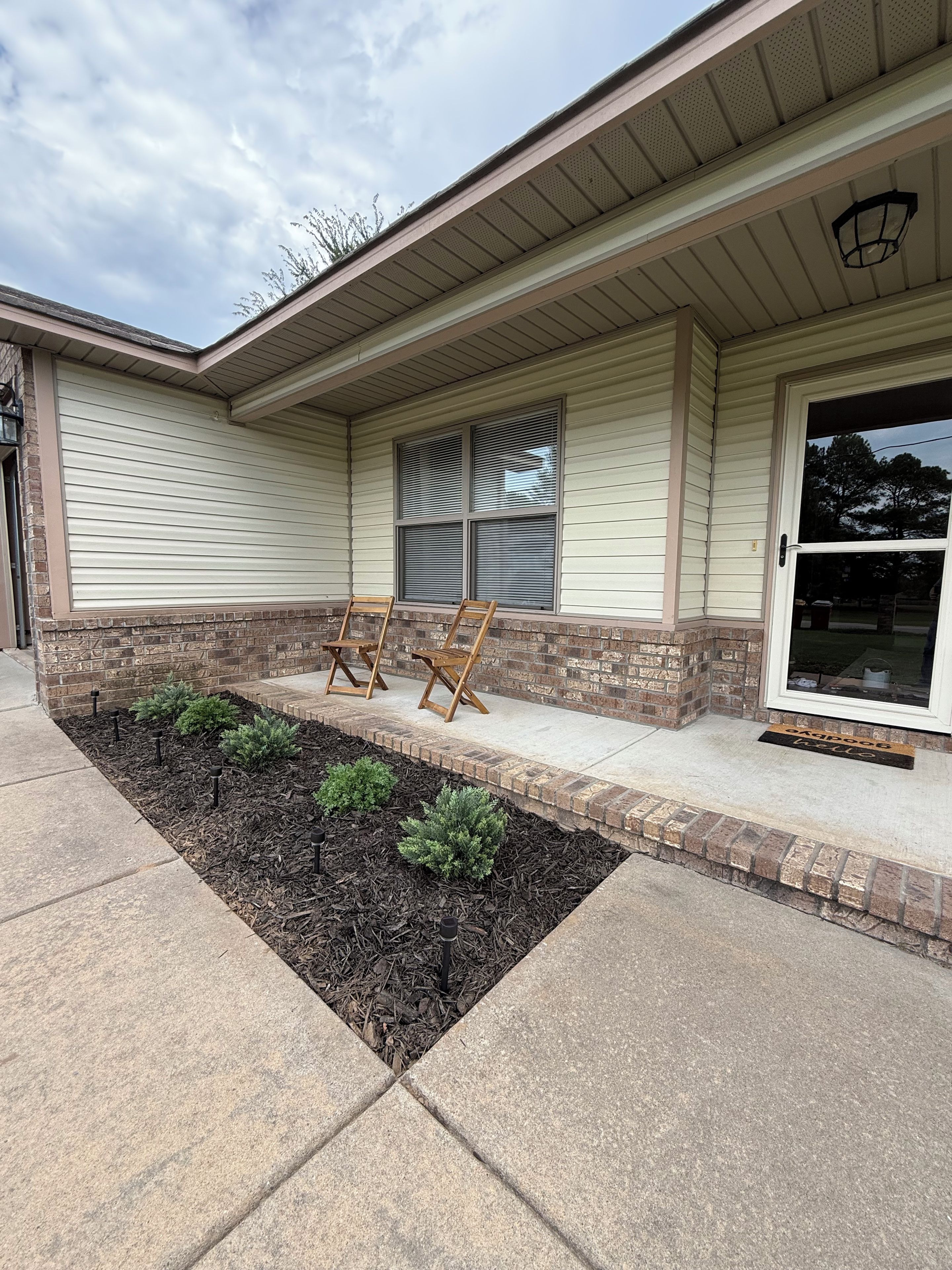 A house with a covered porch, a brick foundation, and a small garden area with mulch and plants.