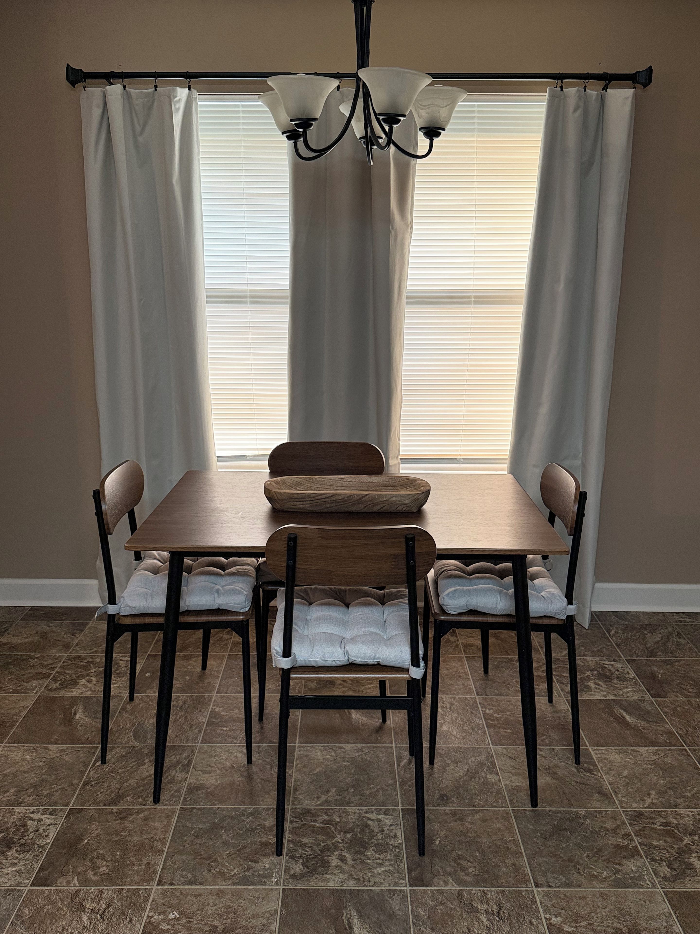 A dining area with a table and four chairs, a chandelier, and a window with blinds.