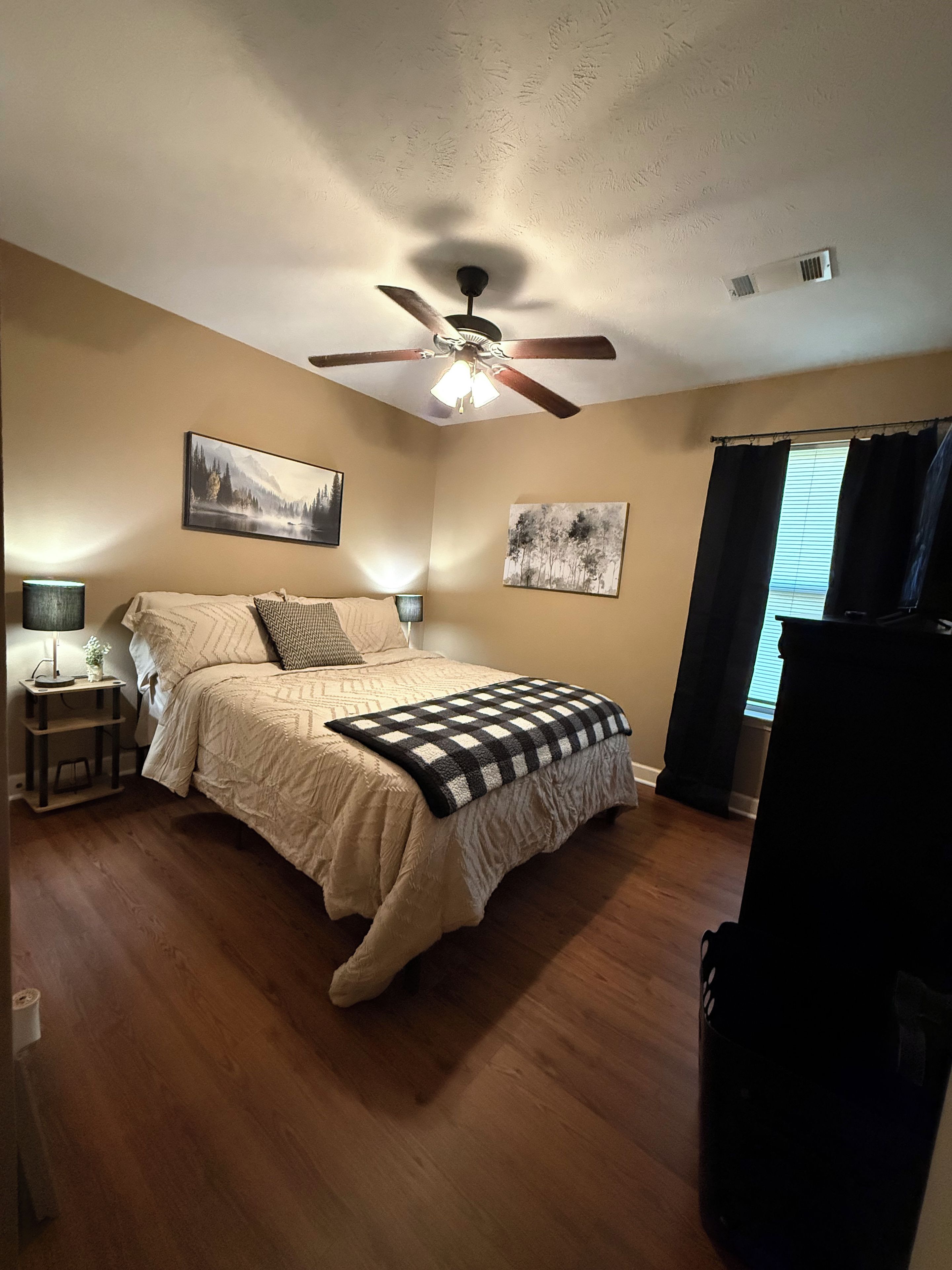 A bedroom with a bed, ceiling fan, wall art, and a window with curtains.