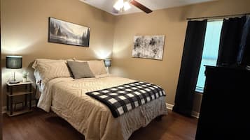 A bedroom with a bed, ceiling fan, wall art, and a window with curtains.