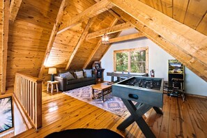 A cozy loft-style room with a wooden ceiling, a sofa, a coffee table, a foosball table, and a window with a view of trees.