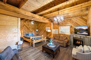 A log cabin interior with a wooden ceiling, a bed, a sofa, a fireplace, and a television.