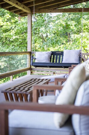 A wooden deck with a swing chair and a table, surrounded by greenery. - Endless Views - Game Room, Arcade, Hot Tub! (Sevierville)