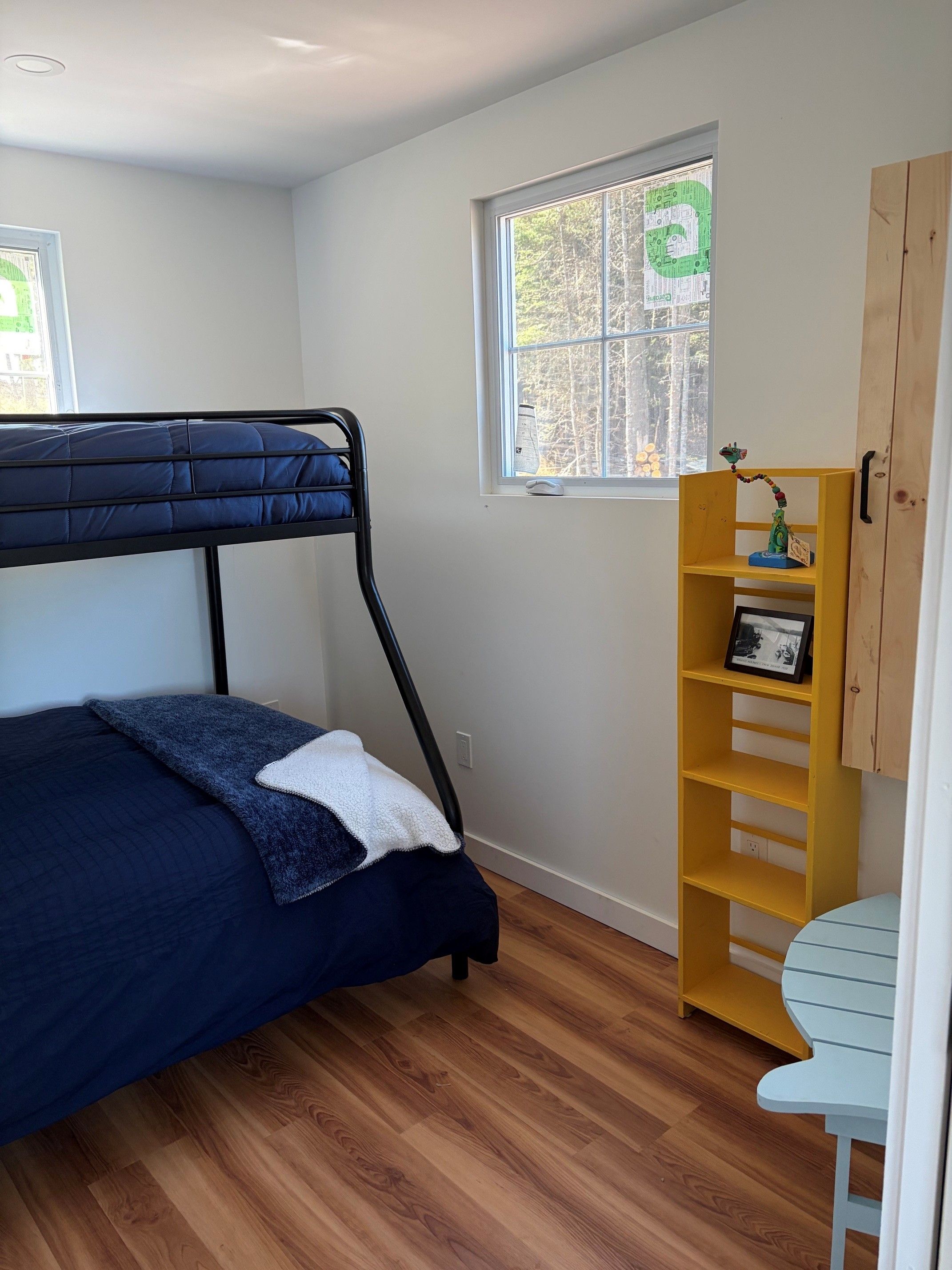 A bedroom with a bunk bed, a wooden floor, a window with a view of trees, and a yellow shelf with a picture frame.
