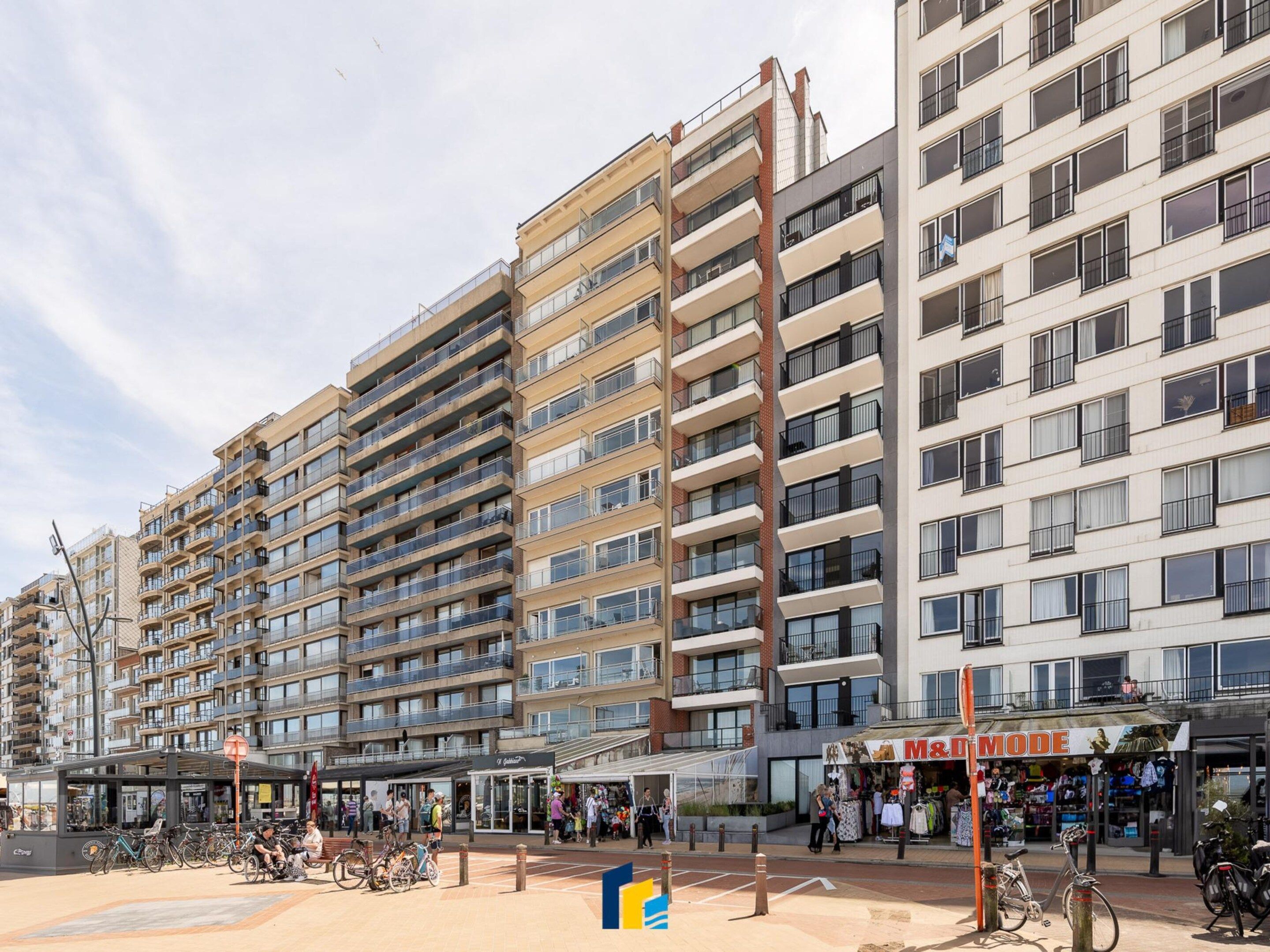 Un immeuble résidentiel à plusieurs étages, doté de balcons, d’un espace extérieur couvert aménagé en coin salon et d’une devanture de magasin avec une enseigne.