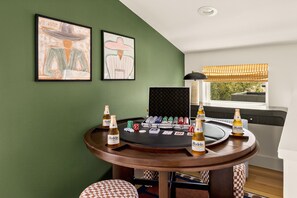 A room with a green wall, two framed artworks, a poker table with chips and bottles, and a window with blinds. - Eastside ATX Social | Golf Sim • Hot Tub • Sauna (Austin)