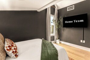 A modern bedroom with a bed, a television mounted on the wall, and a window with curtains. - Eastside ATX Social | Golf Sim • Hot Tub • Sauna (Austin)