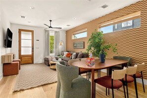 A modern living room with a sofa, coffee table, and chairs, a dining area with a table and chairs, a ceiling fan, and a television mounted on the wall. - Eastside ATX Social | Golf Sim • Hot Tub • Sauna (Austin)