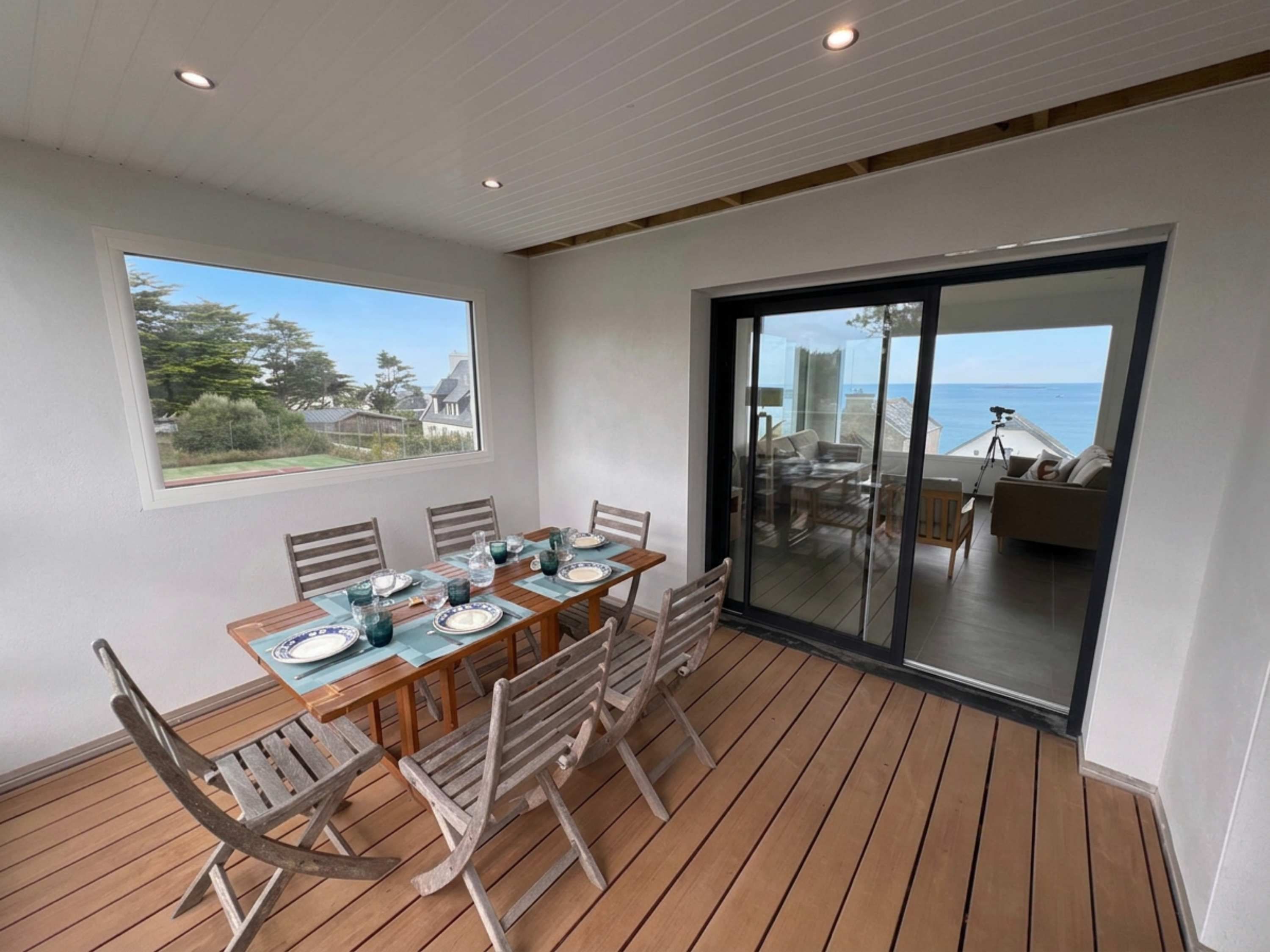 Un espacio de comedor al aire libre cubierto con una mesa y sillas de madera, vista al mar a través de un ventanal y un balcón con mobiliario exterior.