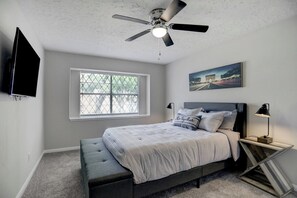 A bedroom with a bed, a ceiling fan, a window, a wall-mounted TV, and a bedside table with a lamp. - Home away from Home! (Spring)