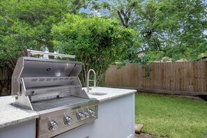 A portable gas grill with a sink and four burners, set in a backyard with a wooden fence and trees. - Home away from Home! (Spring)