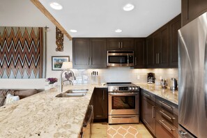 A modern kitchen with dark cabinets, stainless steel appliances, a granite countertop, and a decorative wall piece. - Snowmass Retreat: Vaulted Ceilings, Wood FP, Balcony W/ Grill & Great Views, W/D, Parking, Pool & HT (Snowmass Village)