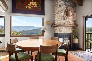 A dining area with a round wooden table and wicker chairs, a stone fireplace, and a view of mountains through large windows. - Snowmass Retreat: Vaulted Ceilings, Wood FP, Balcony W/ Grill & Great Views, W/D, Parking, Pool & HT (Snowmass Village)