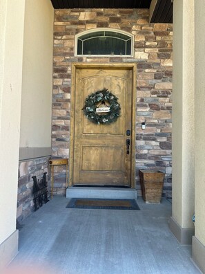 A wooden front door with a welcome wreath, a stone wall, a small wooden bench, and a trash bin.