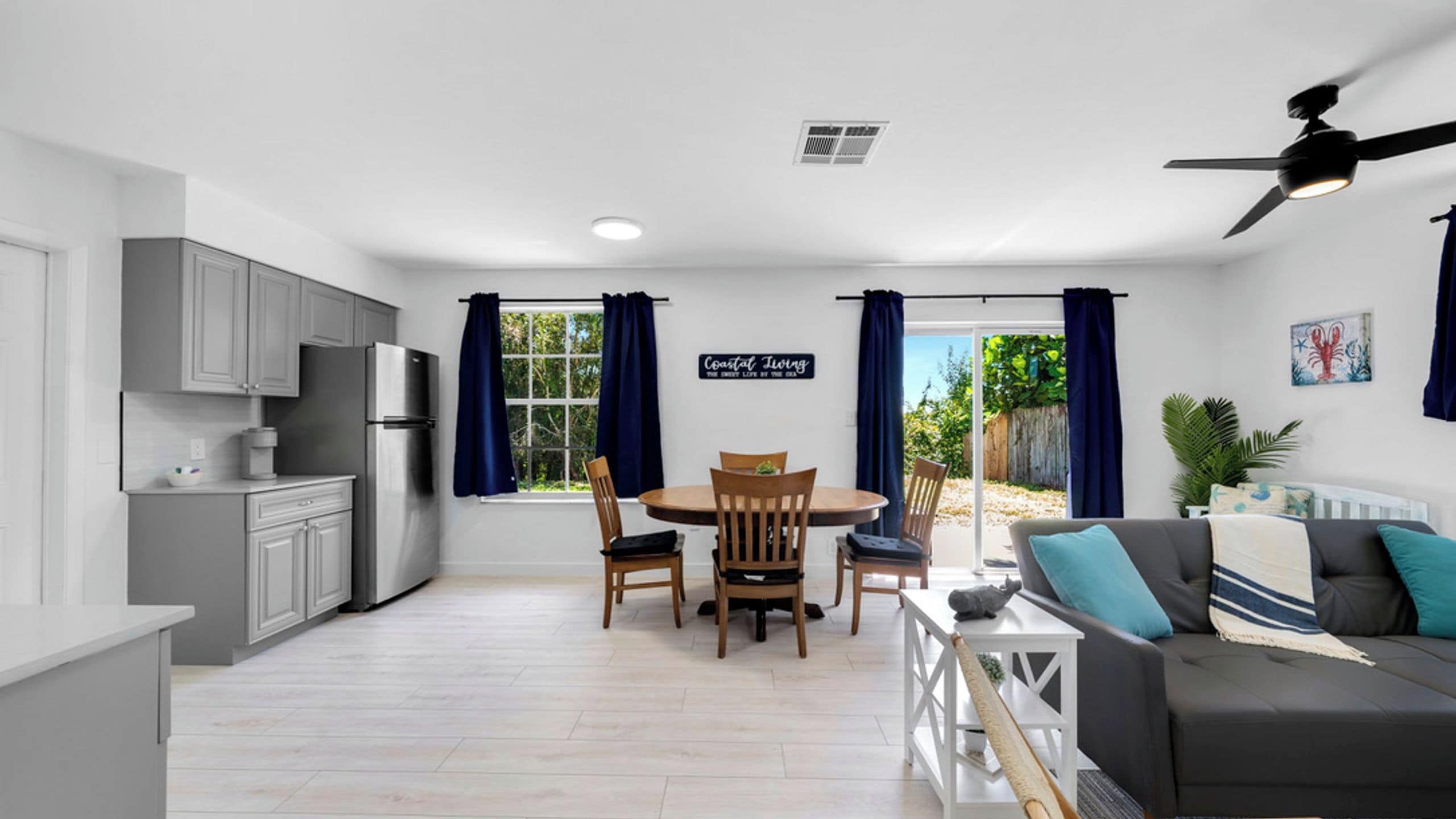 Una cocina moderna con armarios grises y un refrigerador empotrado, un comedor con mesa y sillas redondas, y una sala de estar con sofá gris y ventilador de techo.