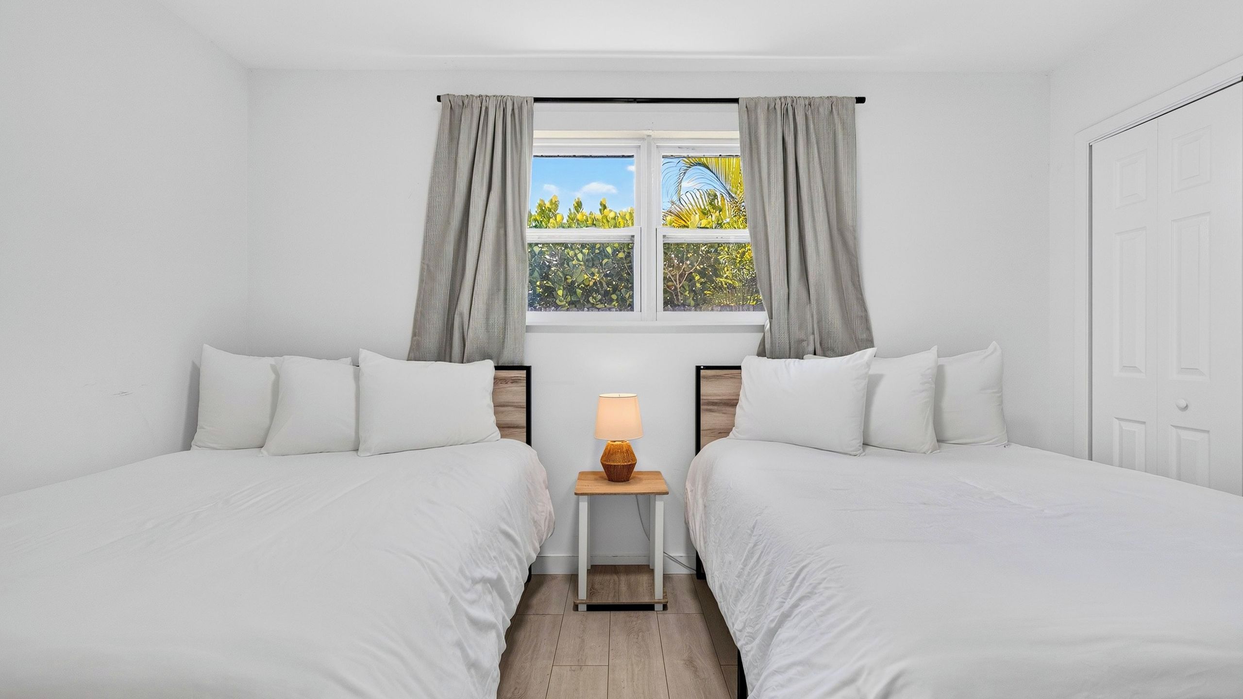 Quarto de hotel com duas camas, uma janela com cortinas, mesa de cabeceira com abajur e vista para o verde lá fora.