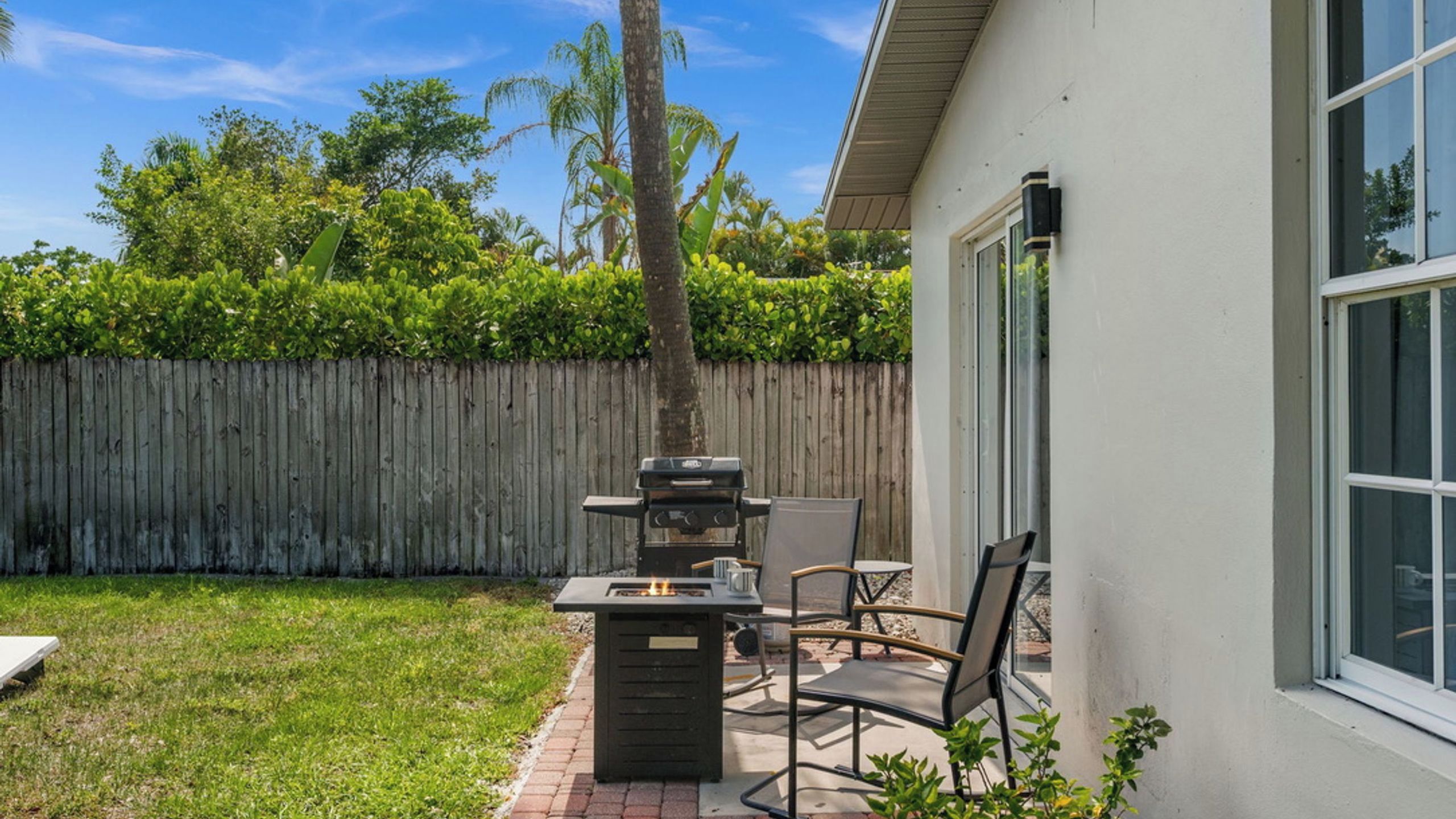 Un patio con una parrilla y sillas, una cerca de madera, y una casa con ventana.