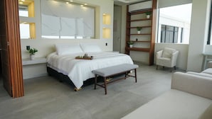 A modern bedroom with a bed, bedside tables, a bench, a chair, and a shelving unit with plants. - Aurea Vita (Tres Vidas Acapulco Diamante)