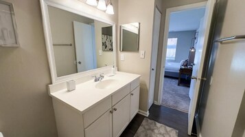 A bathroom with a white vanity, a large mirror, and a door leading to a bedroom.