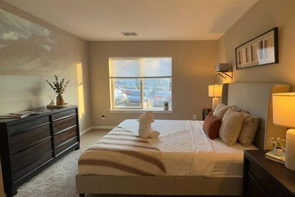 Un dormitorio con cama, lámparas de noche, cómoda, ventana con vista a coches estacionados y un cuadro en la pared.