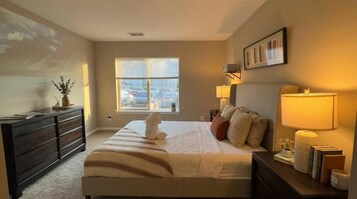 A bedroom with a bed, bedside lamps, a dresser, a window with a view of parked cars, and a painting on the wall.