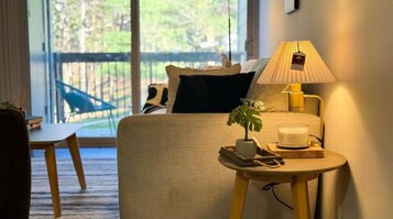Habitación de hotel con cama, mesita de noche con lámpara, una planta pequeña y una vela. Se observa una silla y vista al exterior a través de la ventana.