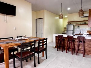 Un coin repas avec une table et des chaises en bois, une cuisine équipée d’un réfrigérateur et d’un évier, et des luminaires suspendus en osier.