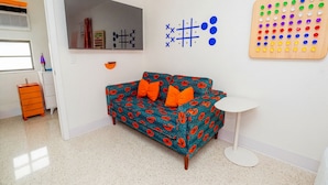 Une pièce comprenant un canapé à motifs, une petite table blanche, un jeu de société fixé au mur et un meuble de rangement.