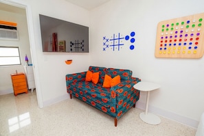 Une pièce comprenant un canapé à motifs, une petite table blanche, un jeu de société fixé au mur et un meuble de rangement.