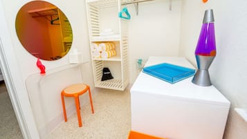 A room with a white wardrobe, a round mirror, a small orange stool, a blue tray, and a lava lamp.
