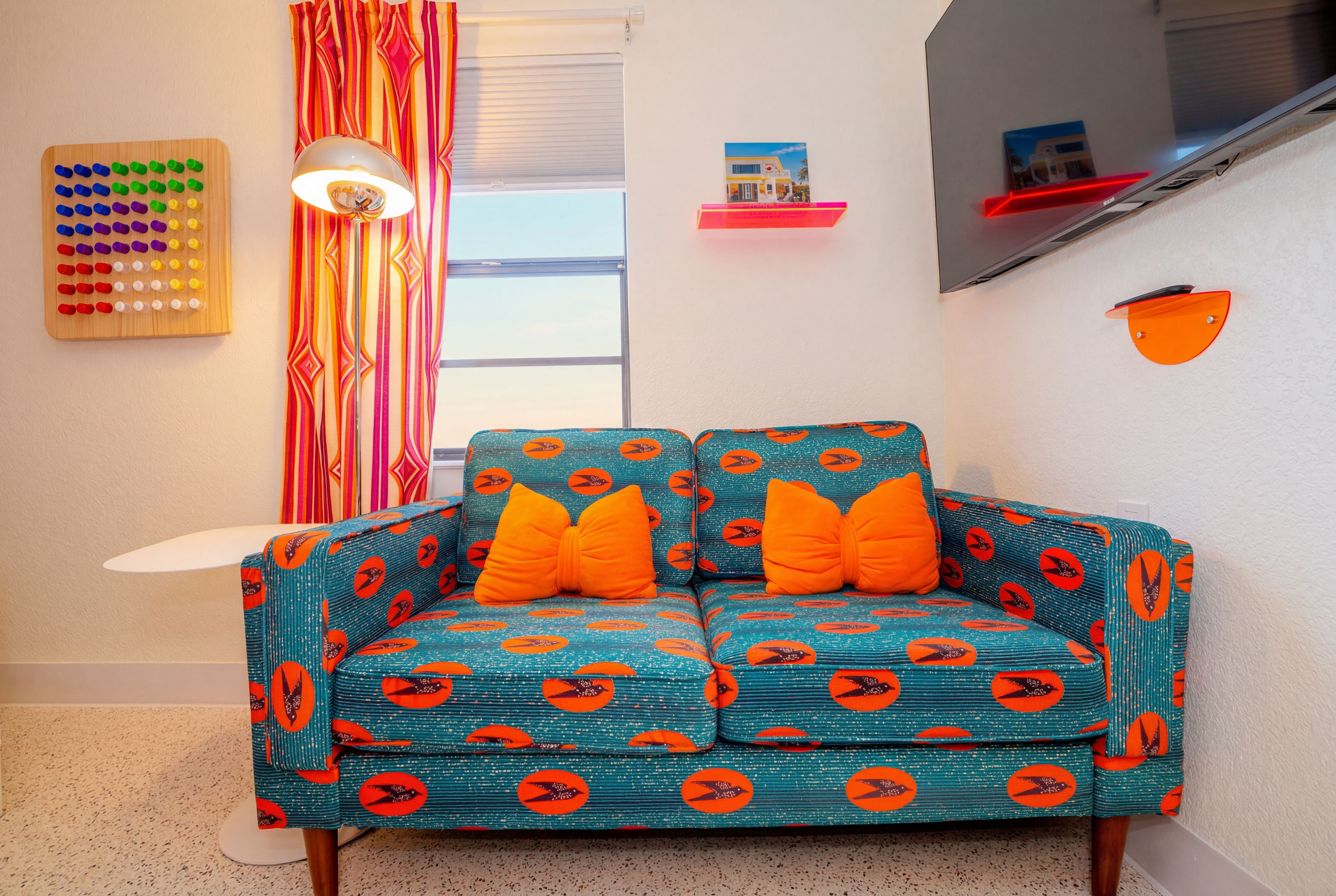 A room with a teal sofa with orange cushions, a wooden bead board, a striped curtain, a lamp, a small shelf, and a wall-mounted TV.