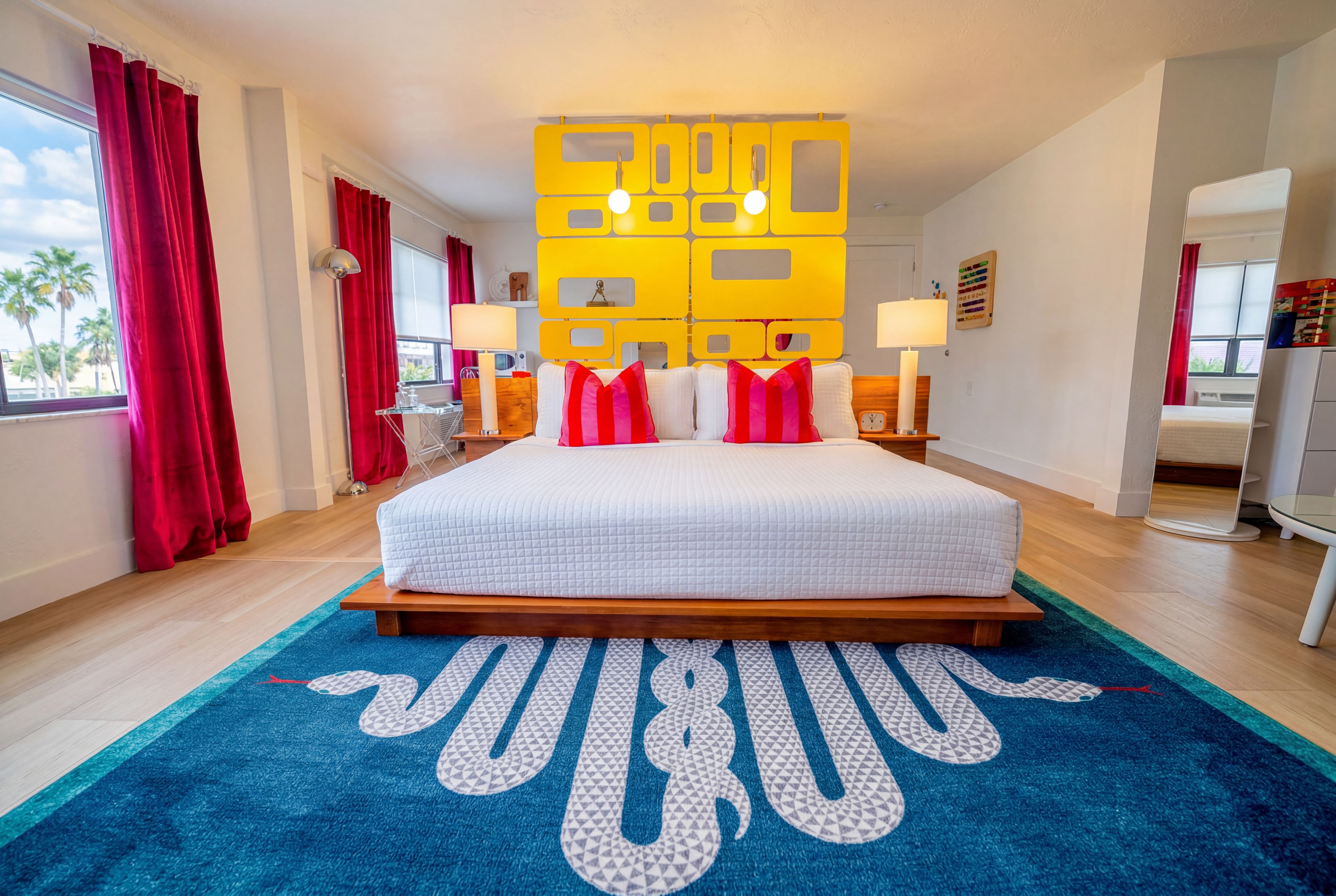 Una habitación de hotel con una cama grande, cortinas rojas, un tapete azul y un adorno amarillo en la pared.